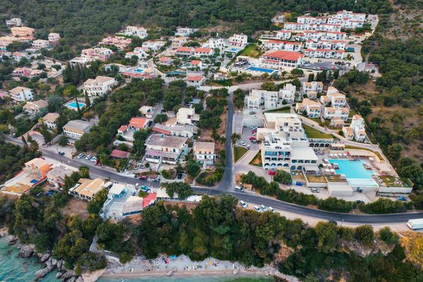 Aerial view of a coastal Greek village with houses, roads, and a hotel with a pool.